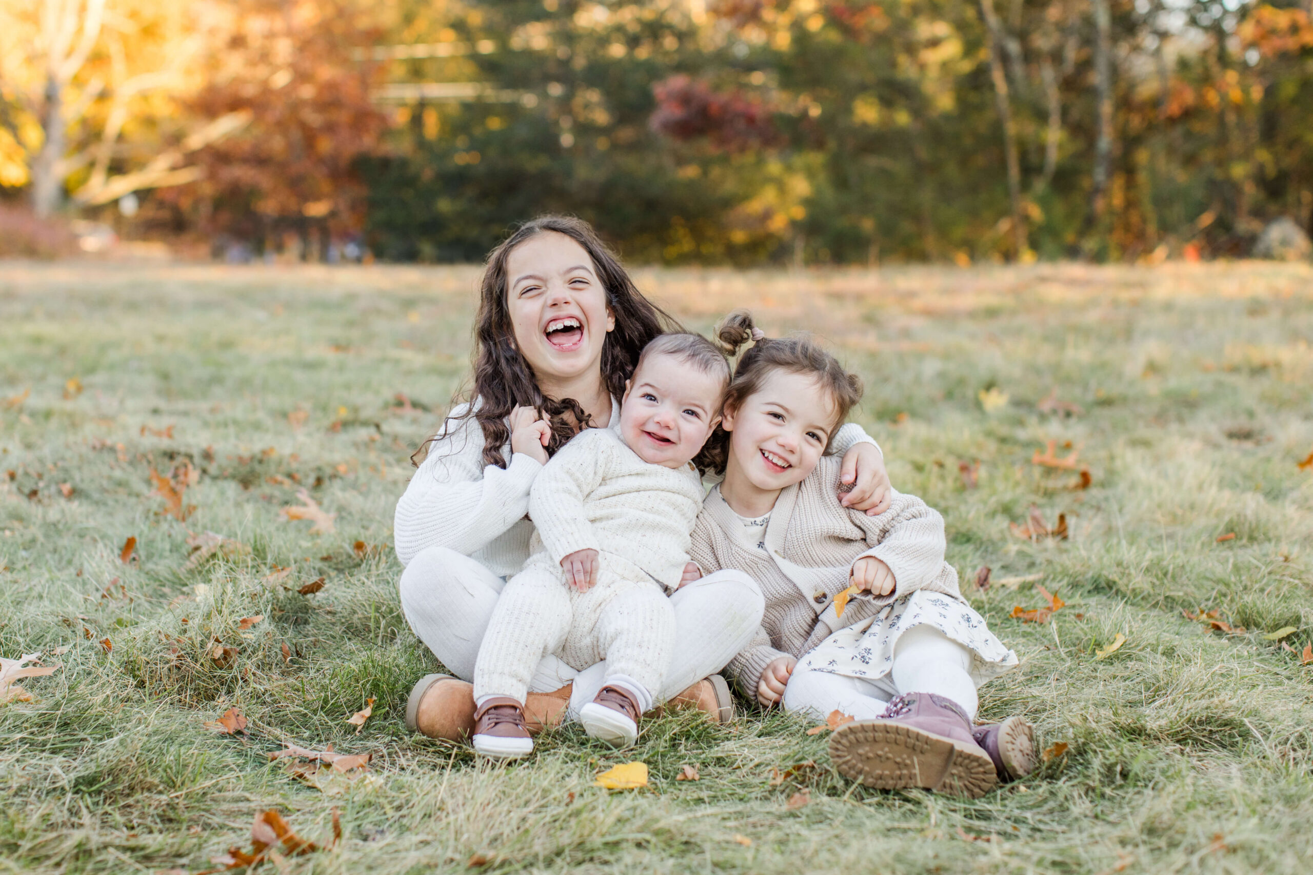 Three young siblings sitting together and laughing during a fall family photo session, surrounded by grass, fallen leaves, and colorful autumn trees.