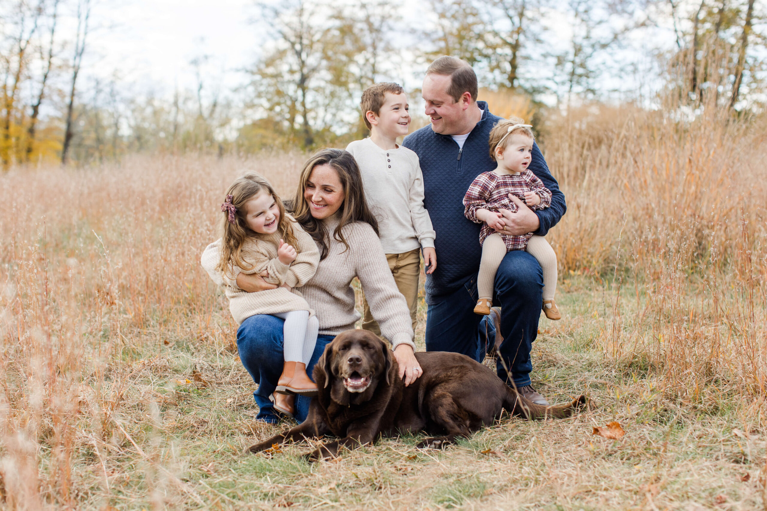 Family of five cuddling together with their chocolate Lab during a relaxed fall mini session in a grassy field.