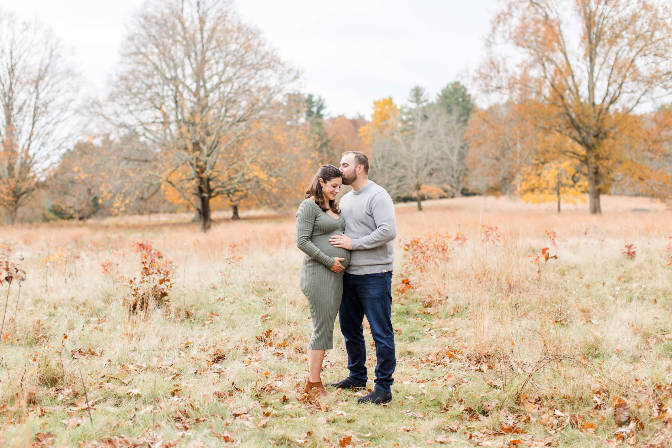 Expecting father kissing his partner’s forehead while resting a hand on her baby bump during a fall maternity photo session.