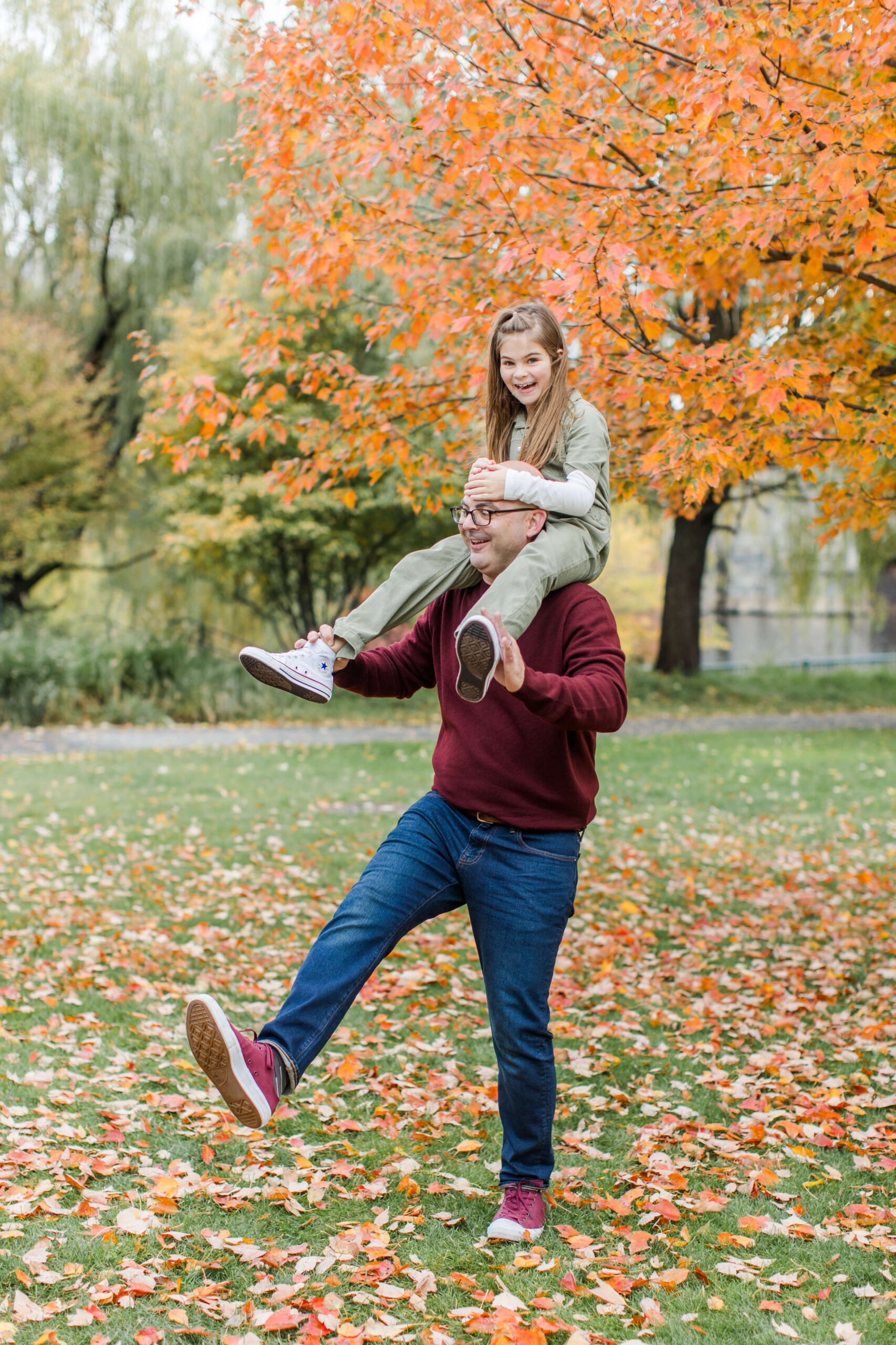 Father carrying his daughter on his shoulders beneath bright orange fall foliage during a family photo session at Boston Public Garden.