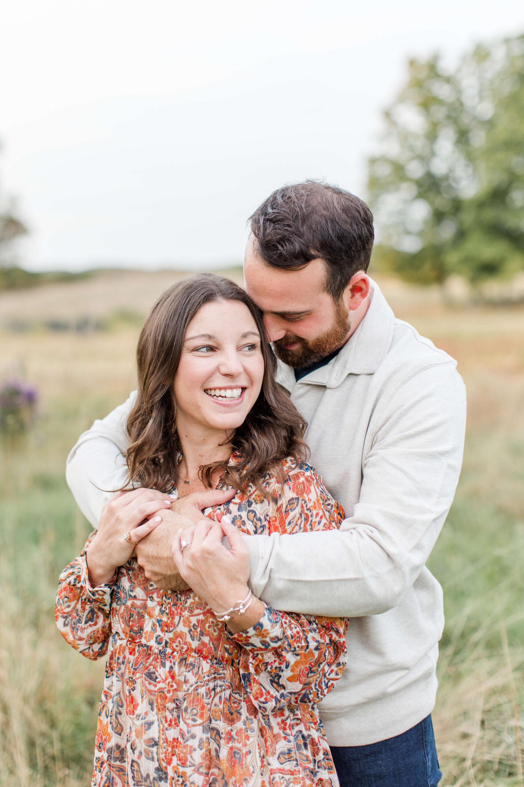 Couple smiling and cuddling together during a fall photo session outdoors, surrounded by soft greenery and autumn scenery.