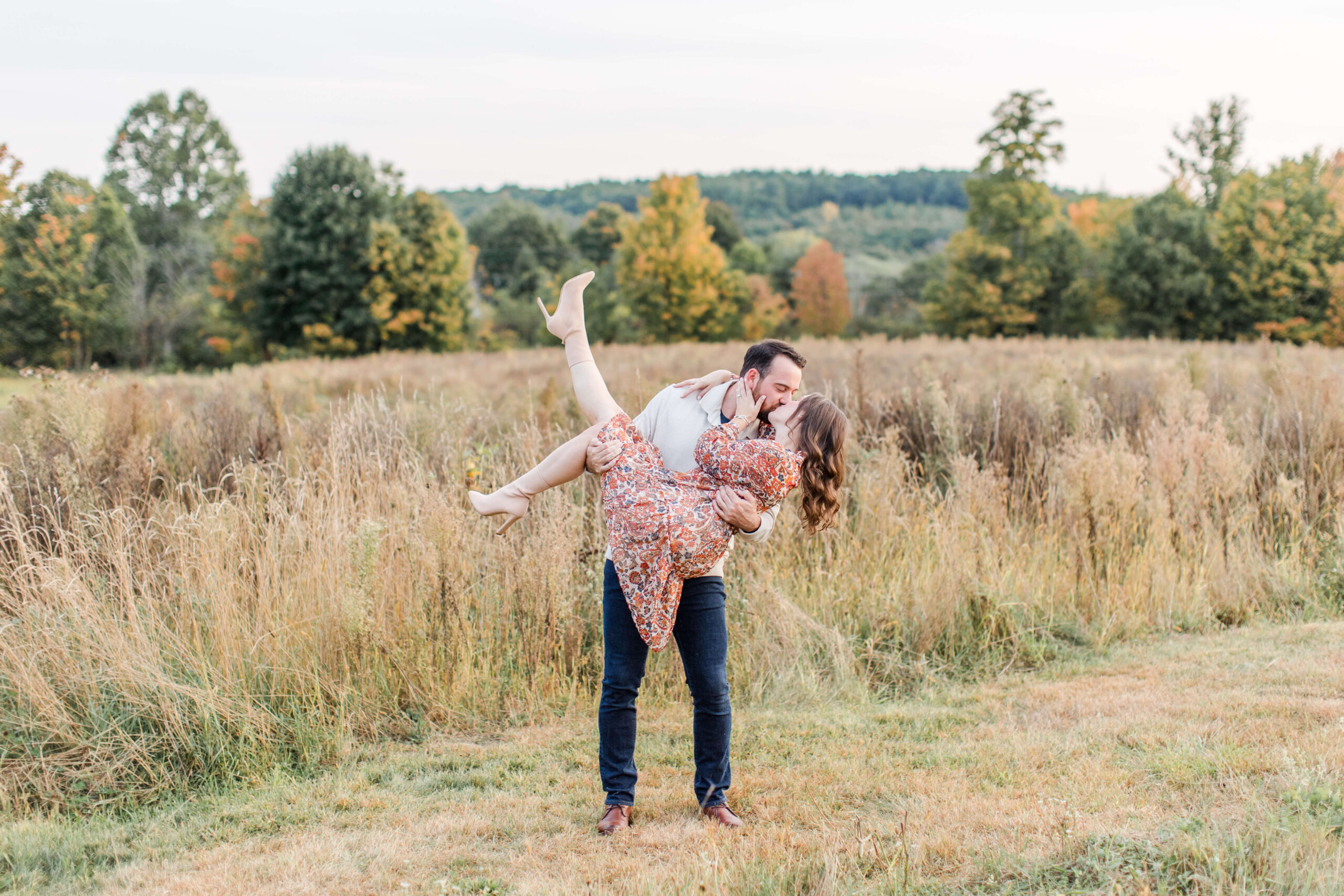 Couple kissing in a grassy field surrounded by autumn trees during a fall photo session.