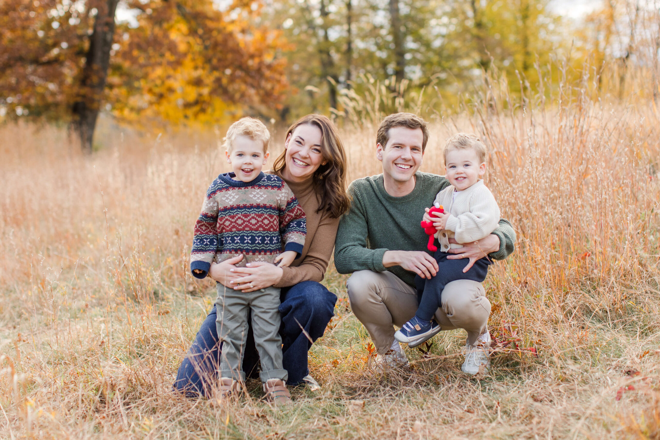 Family of four smiling together in a grassy field surrounded by golden fall foliage during an autumn mini session.
