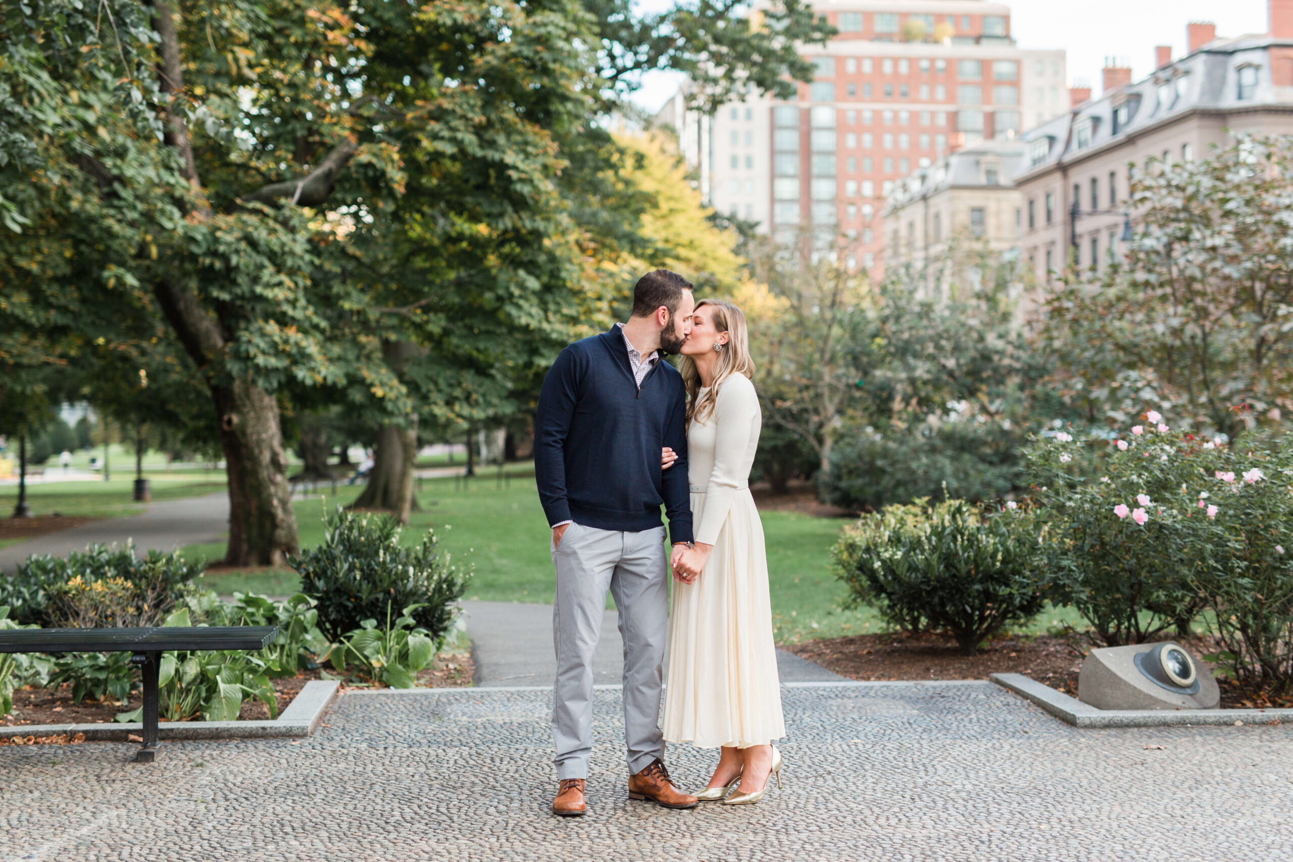 Couple kissing while holding hands during a fall mini session at Boston Public Garden, surrounded by trees, flowers, and city buildings.