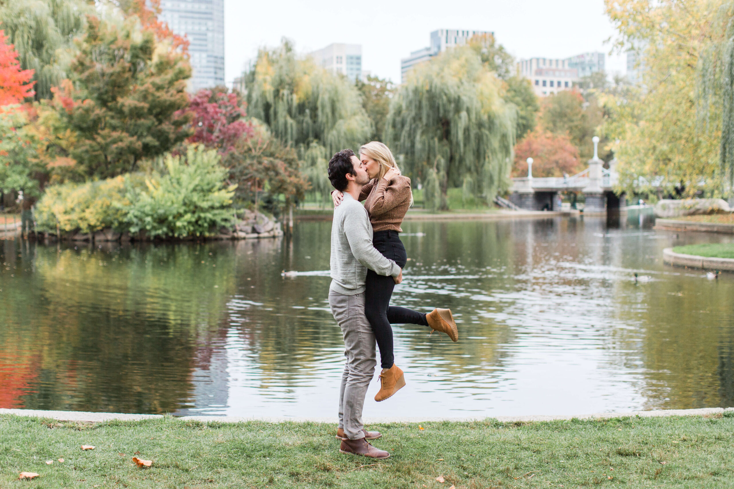 Couple kissing beside the lagoon during a fall mini session at Boston Public Garden, with autumn trees and city buildings in the background.