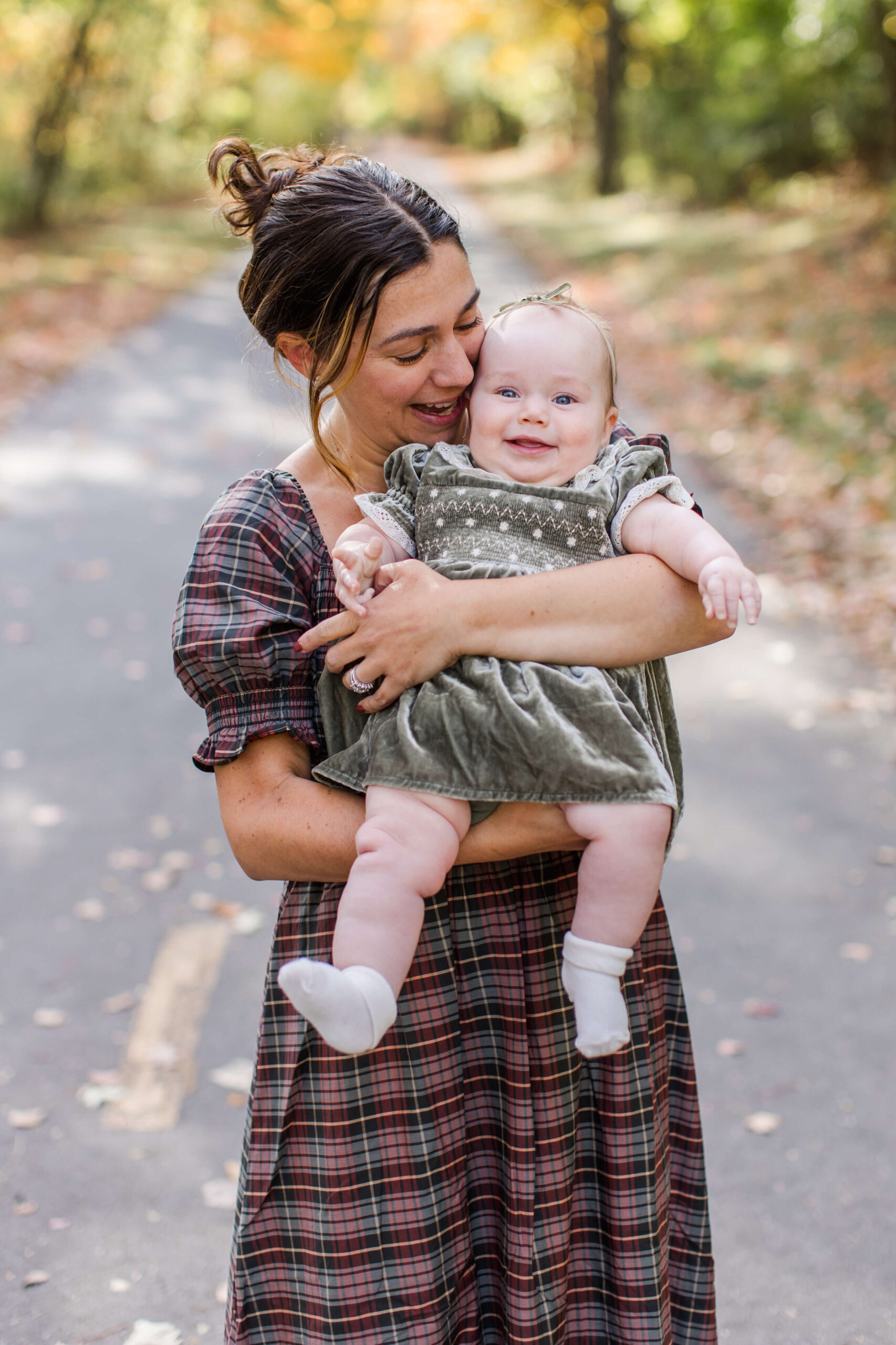 Mother smiling down at her baby while holding her on a tree-lined path during a fall family photo shoot.