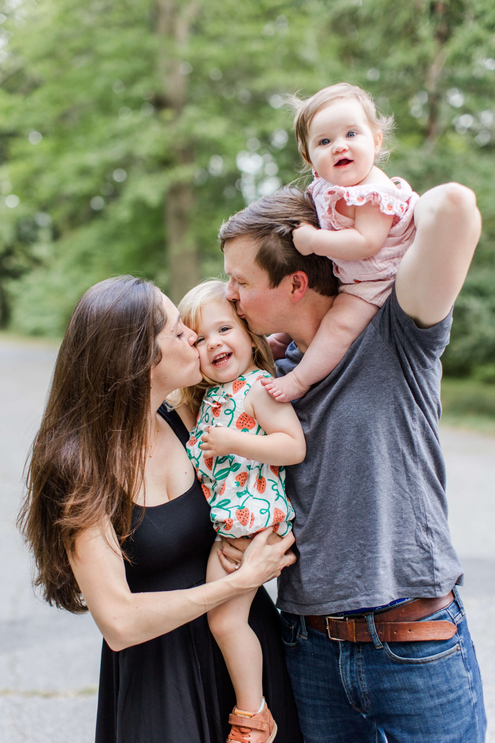 Parents kissing their young daughter while their baby sits on the father’s shoulders during a playful family photo shoot.