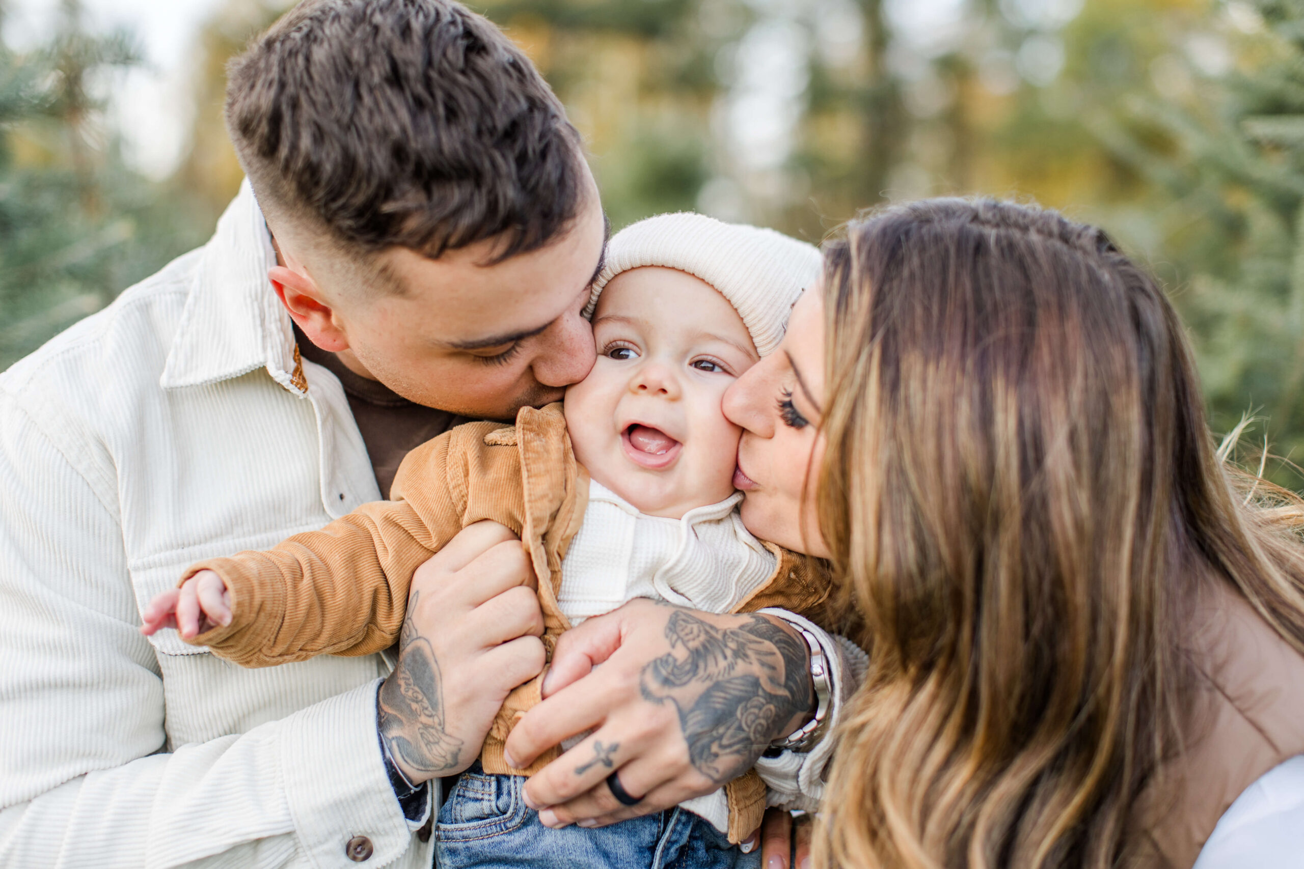 Parents kissing their smiling baby on both cheeks during an outdoor family photo shoot.