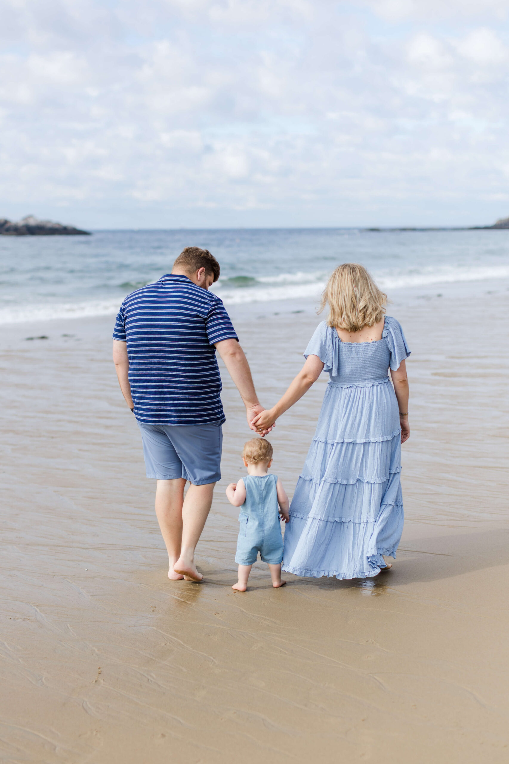 Parents holding hands with their toddler as they walk barefoot along the shoreline during a family beach photo shoot.