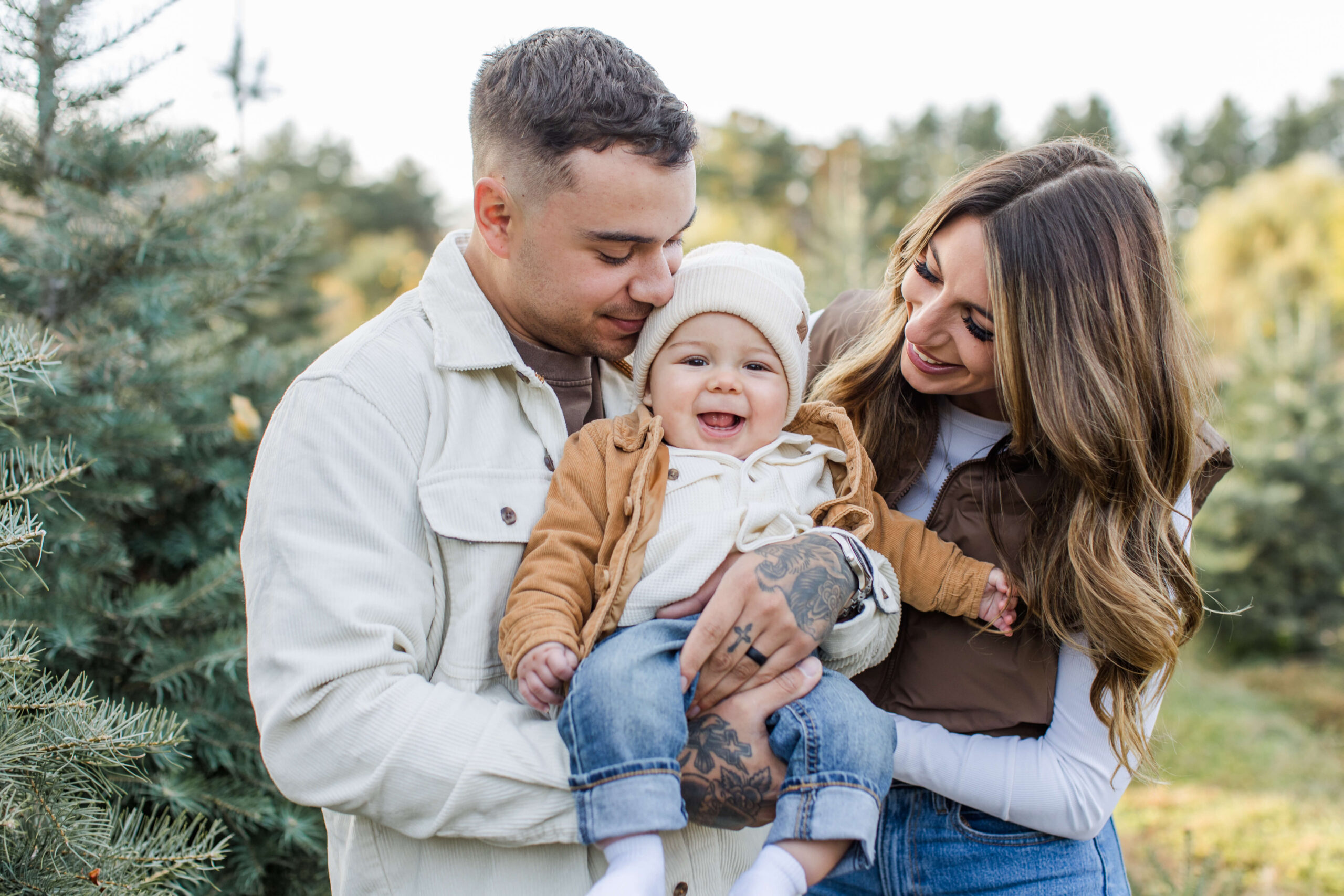 Parents smiling down at their baby during a Christmas mini session surrounded by evergreen trees.