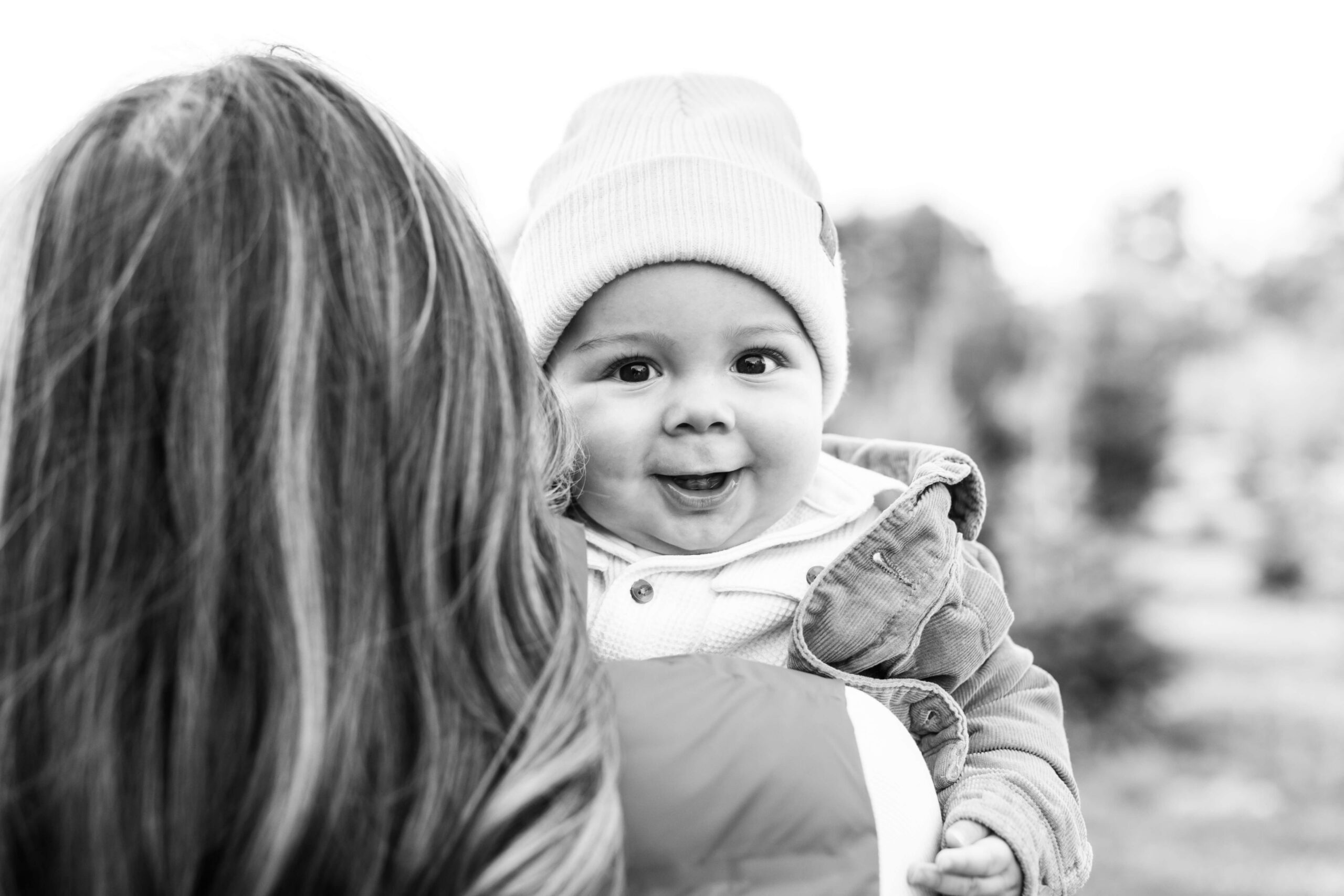 Black and white close-up of a smiling baby in a knit hat being held during a Christmas tree farm photo session.