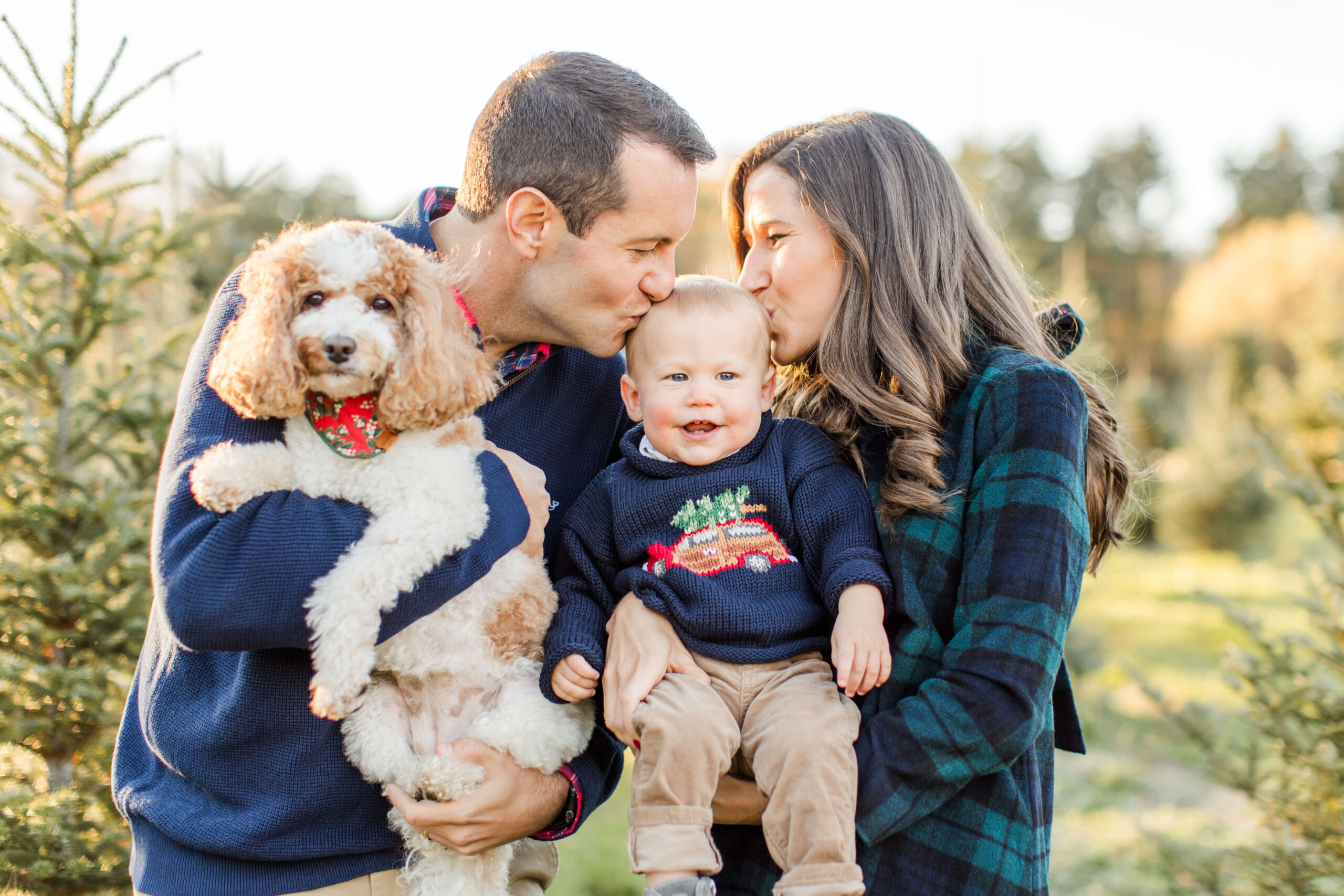 Parents kissing their smiling baby while holding their dog during a Christmas mini session at a Christmas tree farm.