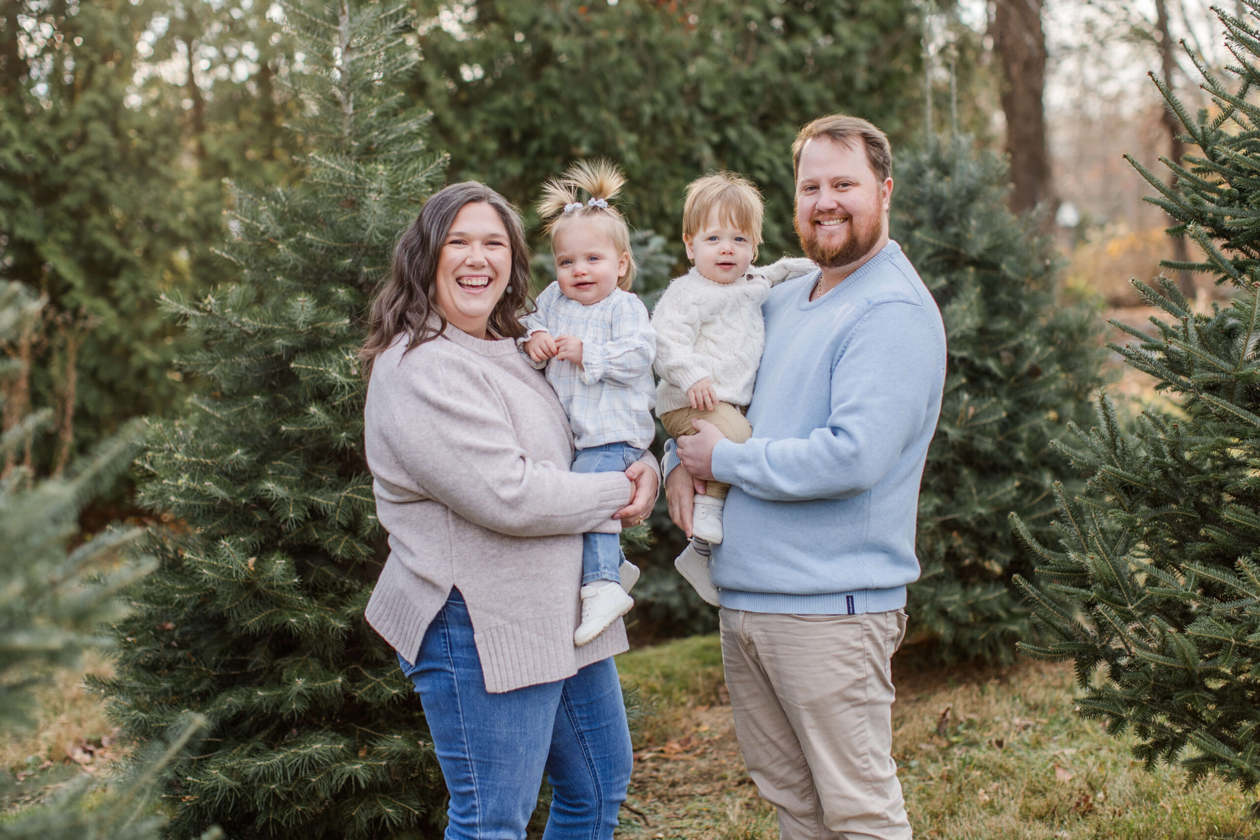 Smiling family of four posing together between evergreen trees during a Christmas mini session at a tree farm.