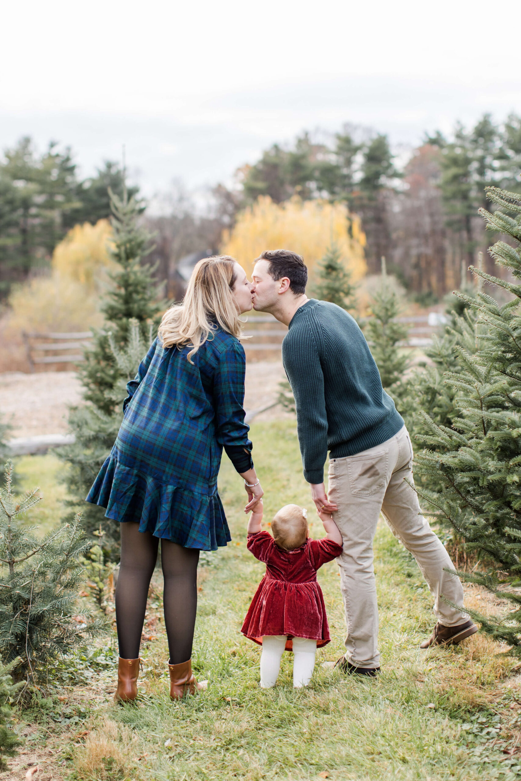 Parents kissing while holding their young daughter’s hands between rows of Christmas trees during a holiday family photo session.